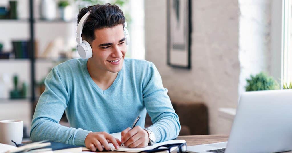 Young man studying online wearing headphones and taking notes